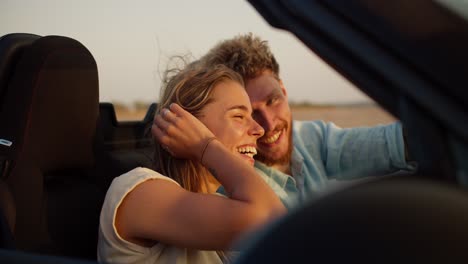 a bearded guy with curly hair in a blue shirt laughs with his blonde girlfriend in a convertible car on a field background