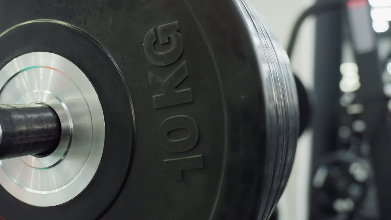 Ten kilogram black bumper plates stacked tightly on chrome barbell sleeve reflect gym lighting while resting on rack, emphasizing weight load, textured rubber surface, engraved kg marking