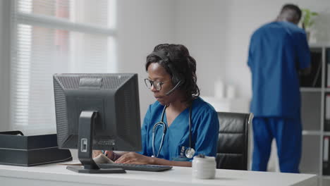 a black woman doctor wearing headphones sits at a table with a computer and takes calls from patients looks at their medical records and enters them into the clinic schedule