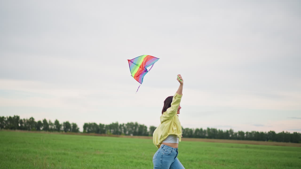 young woman flying kite meadow dusk, rainbow kite arcs across cloudy sky, arms raised during joyful launch, windrippled jacket and denim against vast green field, distant treeline on horizon