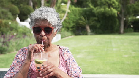 senior african american woman in sunglasses drinking cocktail in garden, copy space, slow motion