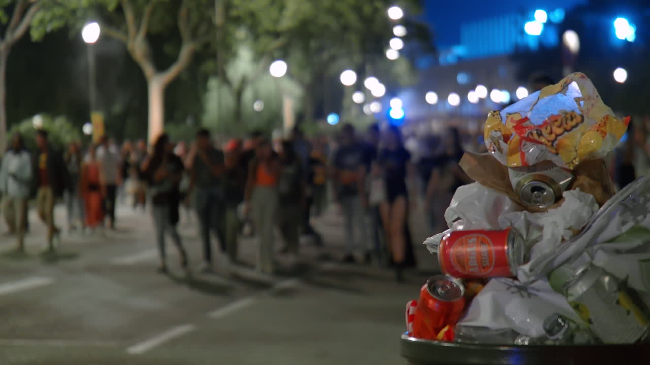 multitud de la vida nocturna caminando más allá de un contenedor de basura lleno de latas y bolsas vacías
