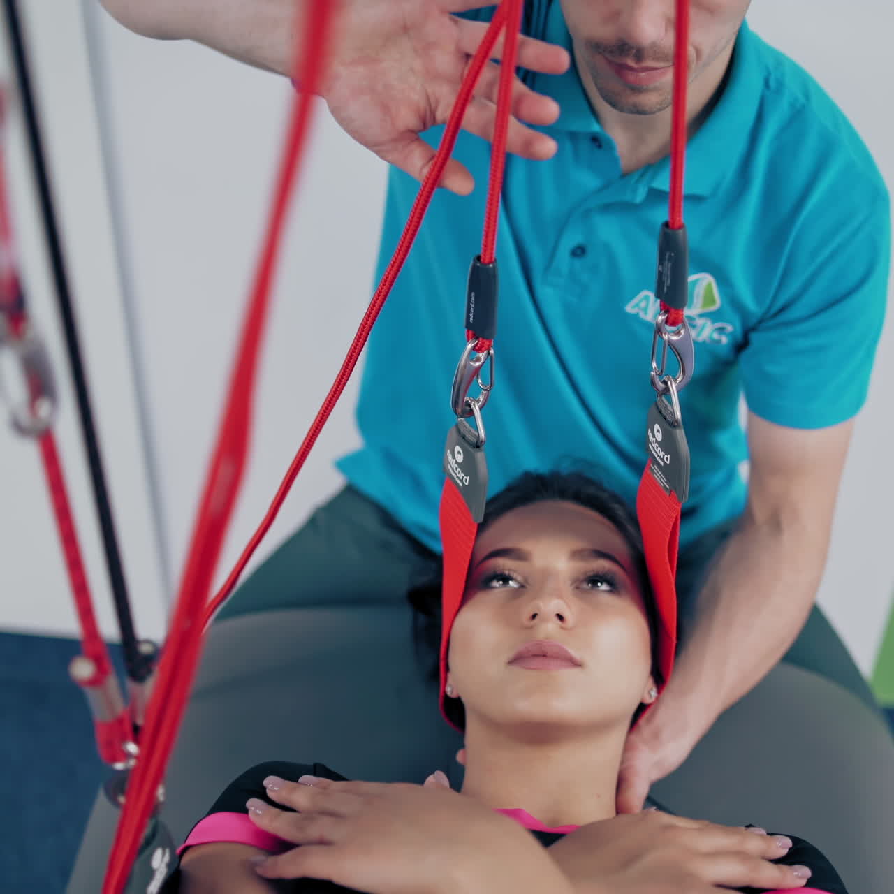 Therapist trainer is supporting female's head in special rope in rehabilitation center. Young woman with her hands on the chest is doing exercises to relieve head pain with personal instructor.