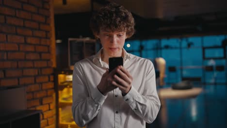 A-young-guy-with-curly-hair-is-pleasantly-surprised-and-jumps-with-happiness-when-he-looks-at-the-screen-of-his-phone-in-the-office