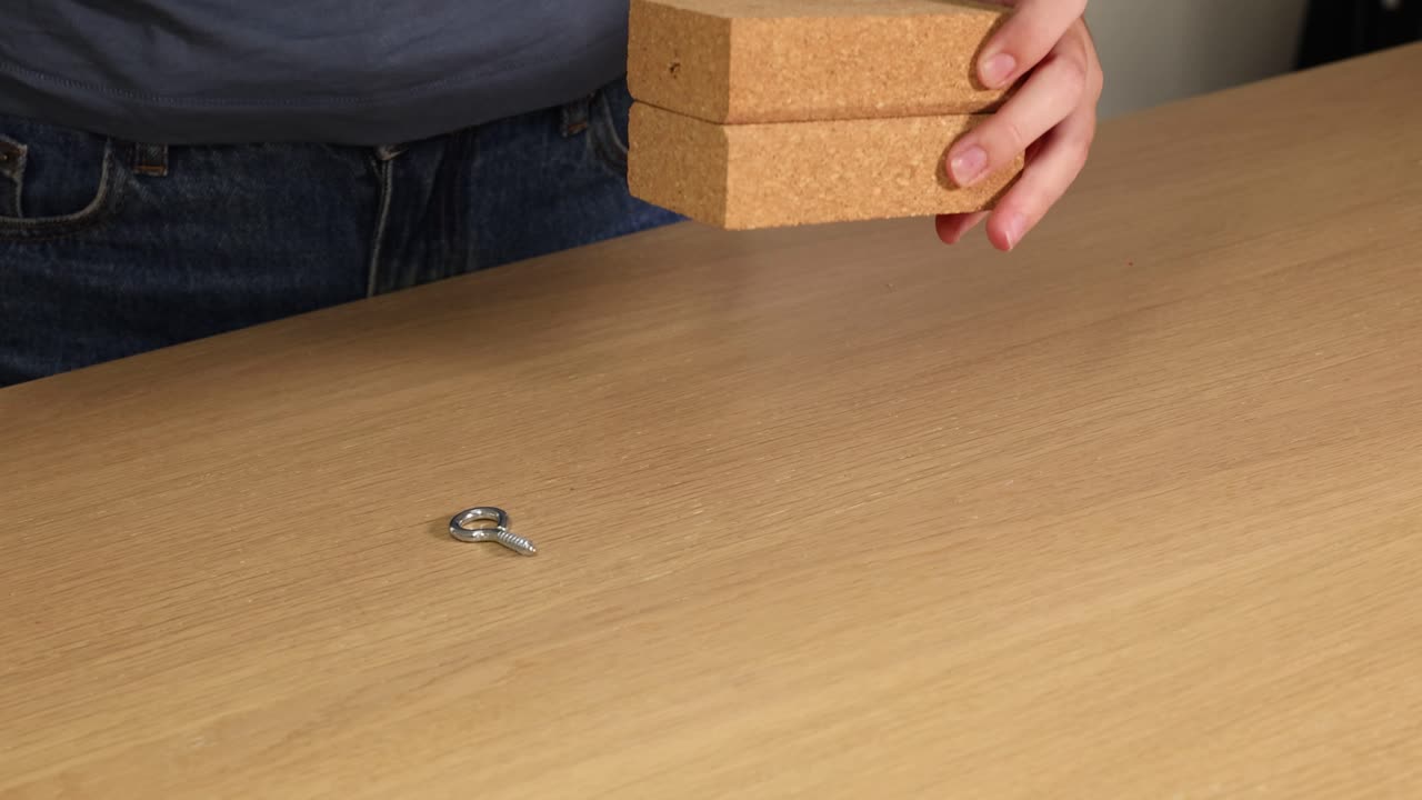 A person inserts a hook screw into a wooden block on a table using hands. Bright lighting, close-up view
