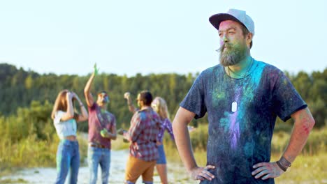 portrait shot of the young attractive sporty man with beard and in a hat looking back at his multiethnic friends who dancing and having fun with paints during holi festival, then turning face to the camera and smiling to the camera