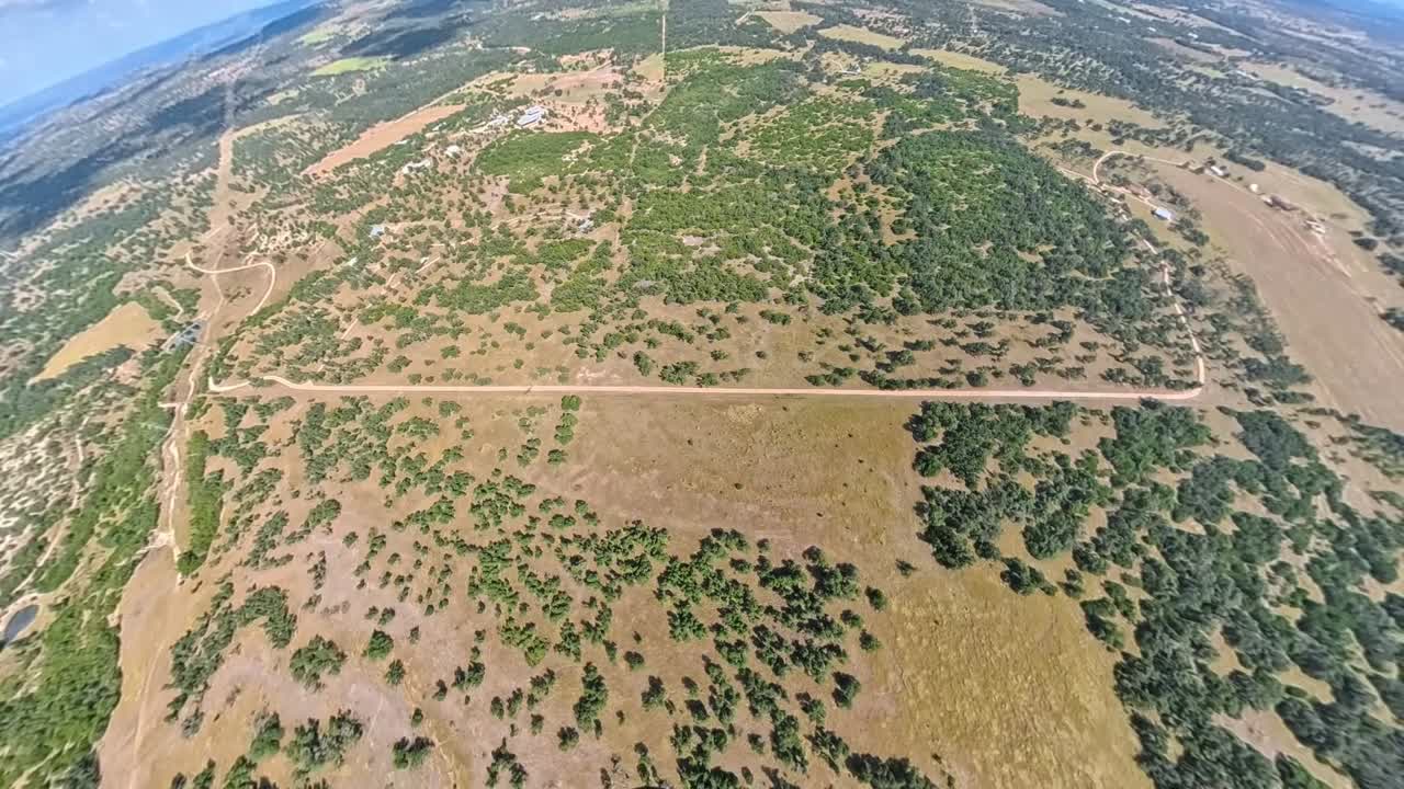 Aerial view from a small yellow airplane flying over dry countryside. The footage captures rugged terrain, scattered trees, dirt roads, and wide open farmland on a sunny day
