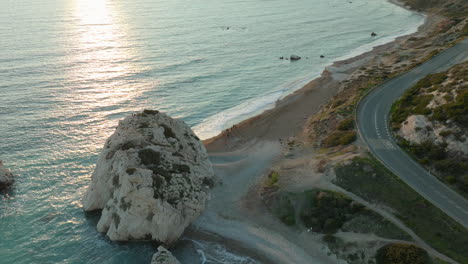 La-Luz-Del-Sol-Dorada-Se-Refleja-En-El-Agua-Cerca-De-La-Icónica-Roca-De-Afrodita,-Con-Una-Sinuosa-Carretera-Costera-Que-Recorre-La-Costa.