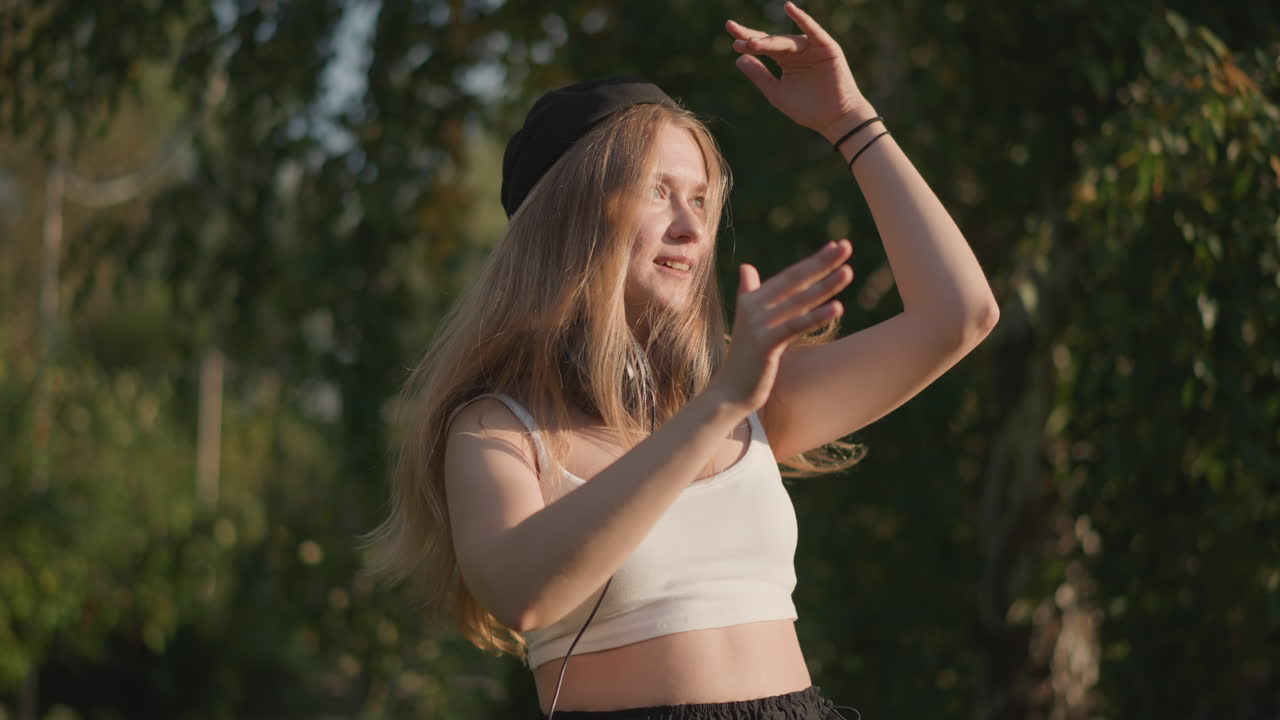 Ung vit kvinna dansar med hörlurar i en solbelyst park, långt hår som fladdrar, klädd i vit crop top och svarta byxor, uttrycksfulla freestyle-rörelser, leende och snurrande, sladden till hörlurarna som svänger, gyllene timmen