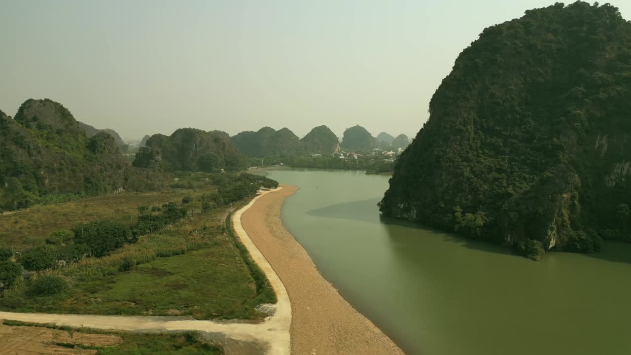 Drone video of a winding river bordered by steep limestone cliffs and a narrow sandy path, surrounded by lush greenery and distant karst hills.