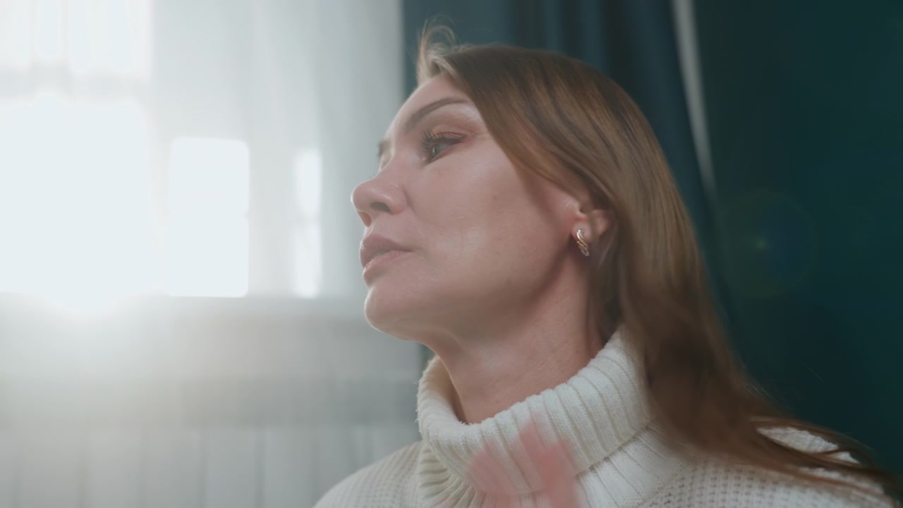 Mature caucasian woman closeup by window, intense light flare and quiet reflection during private coaching session, adjusting hair and listening, soft turtleneck sweater, expressive responses