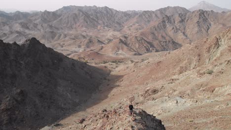 Aerial-epic-shot-from-drone-of-an-young-male-standing-on-top-of-a-rocky-mountain-in-Hatta,-United-Arab-Emirates