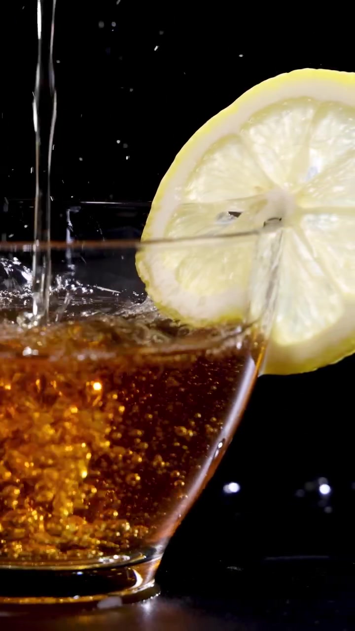 Un getto di tè riempie una tazza di vetro con una fetta di limone, ambientato su uno sfondo scuro.