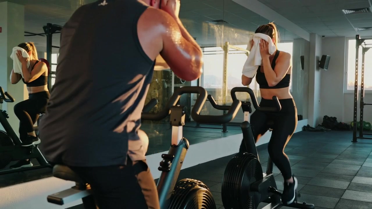 Inizio dell'allenamento, una coppia in abbigliamento sportivo nero pedala su airbike e si asciuga il sudore con asciugamani in palestra.
