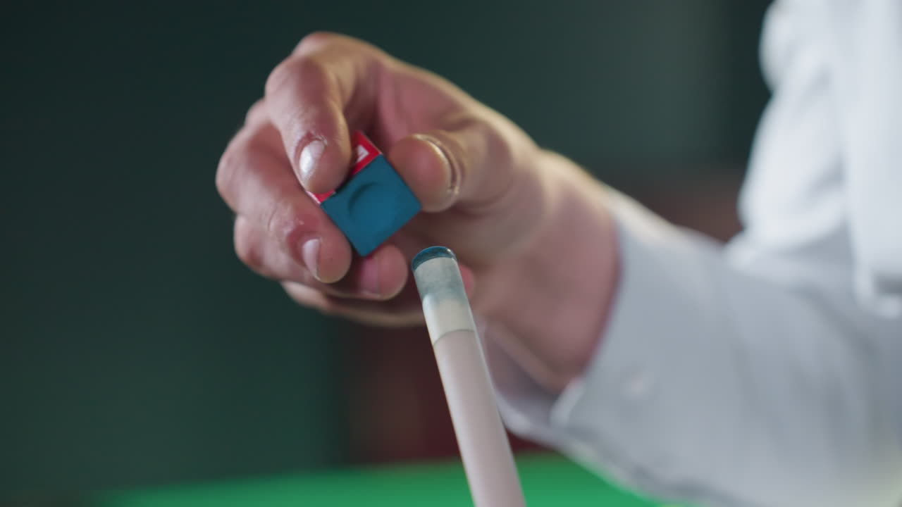 toma de primer plano de la mano con camisa blanca aplicando cuidadosamente la tiza al palo de señal antes de un juego de billar. agarre enfocado en la tiza roja y azul con la mesa verde borrosa en el fondo, destacando la precisión