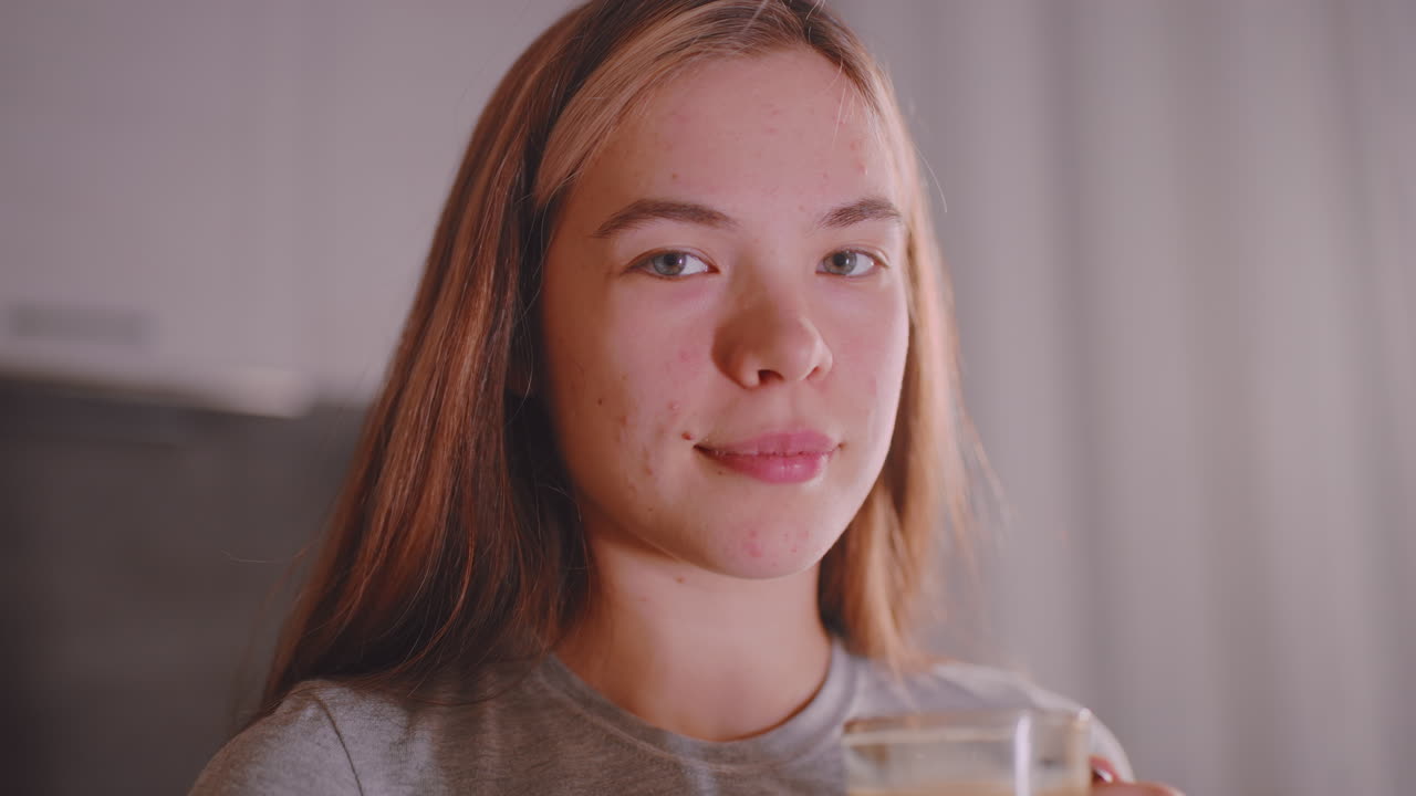 Portrait of young lady holding glass cup with subtle smile and calm gaze, natural indoor lighting highlighting facial features and soft background, capturing everyday lifestyle moment of relaxation