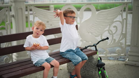 two young boys sit together on a bench, the younger child holds a sausage roll with an expressive look, as if resisting sharing his snack, while the older child stretches