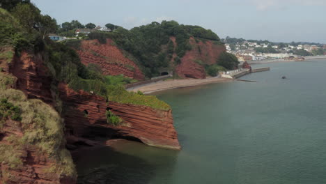 Luftaufnahme,-Die-An-Einem-Sonnigen-Tag-Entlang-Der-Küste-Von-Devon-Mit-Roten-Klippen-Und-Meereshöhlen-Mit-Der-Düsteren-Stadt-In-Der-Ferne-Fliegt