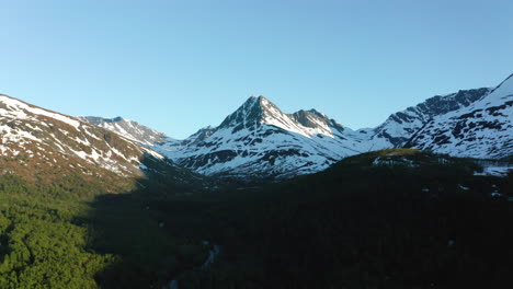 Aerial-view-over-a-valley,-forest-and-a-river,-snowy-Stortinden-mountain-top-in-the-background,-sunny,-summer-day,-in-Lyngen-alps,-North-Norway---Tracking,-drone-shot