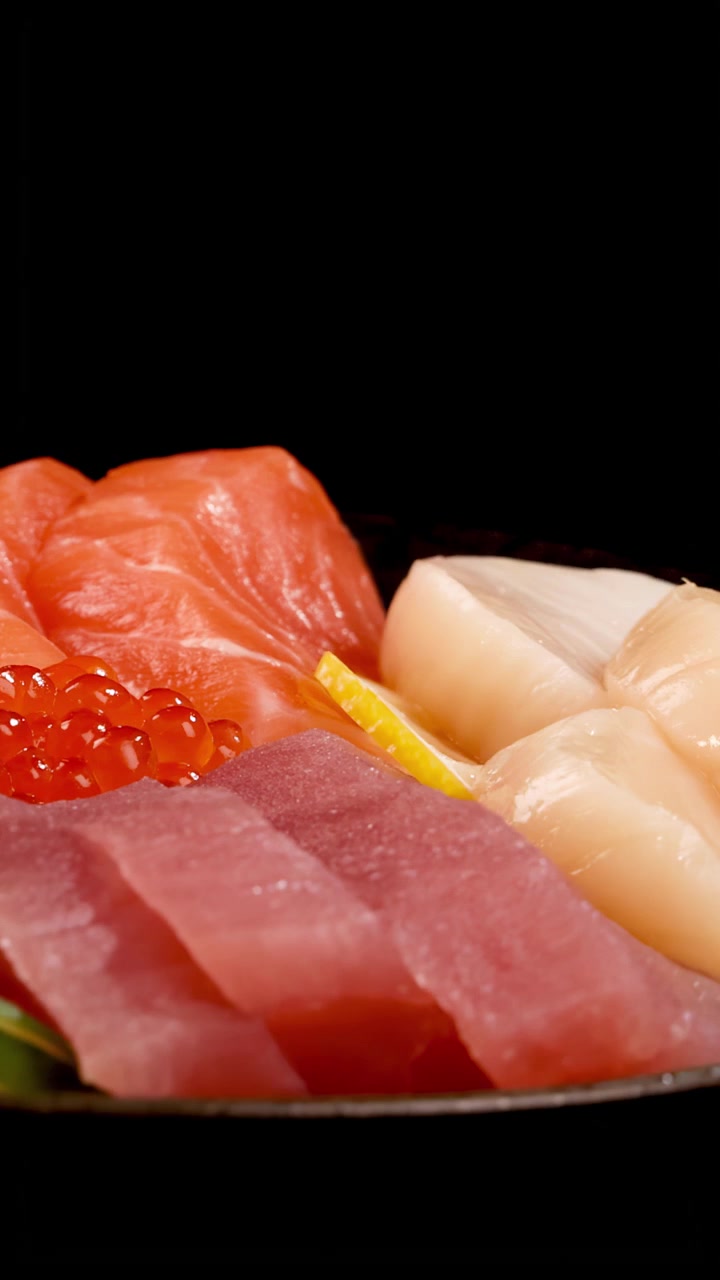 Un vivace piatto di sashimi con salmone, tonno, capasanta, uova di salmone e guarnizioni ruota sotto una brillante illuminazione da studio su uno sfondo nero, evidenziando le texture fresche.