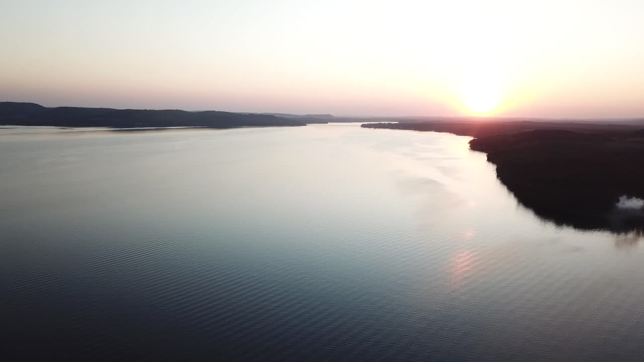 Aerial view of the sun rising over Lake Wisconsin, casting golden light across still waters. Smoke drifts from a wooded shoreline, adding a mystical touch to the peaceful morning landscape.