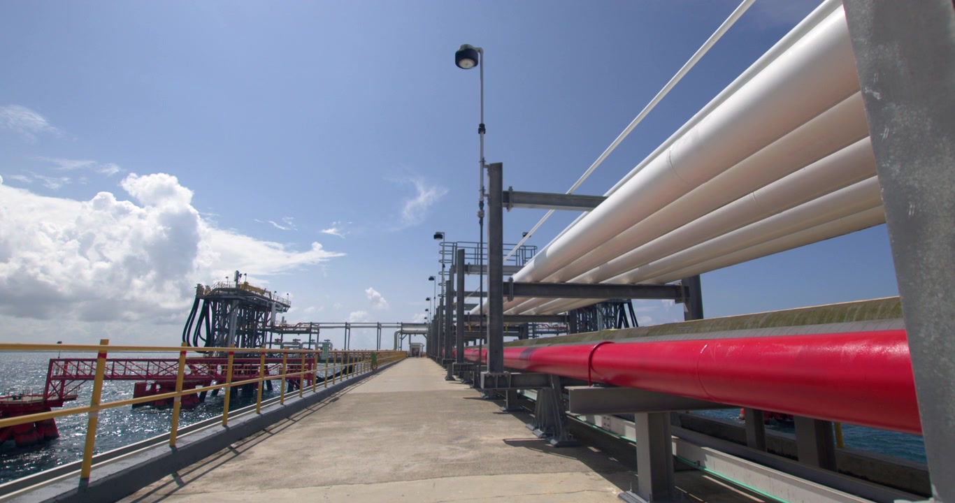 Static wide shot along the pier pathway showing red and white fuel transfer pipelines leading toward the offshore loading platform at Coastal Petroleum terminal in San Pedro de Macoris, Dominican Repu