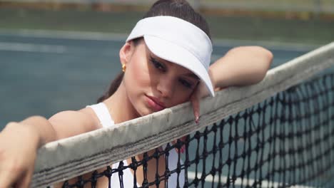 young latina holds her face next to the net at a tennis court