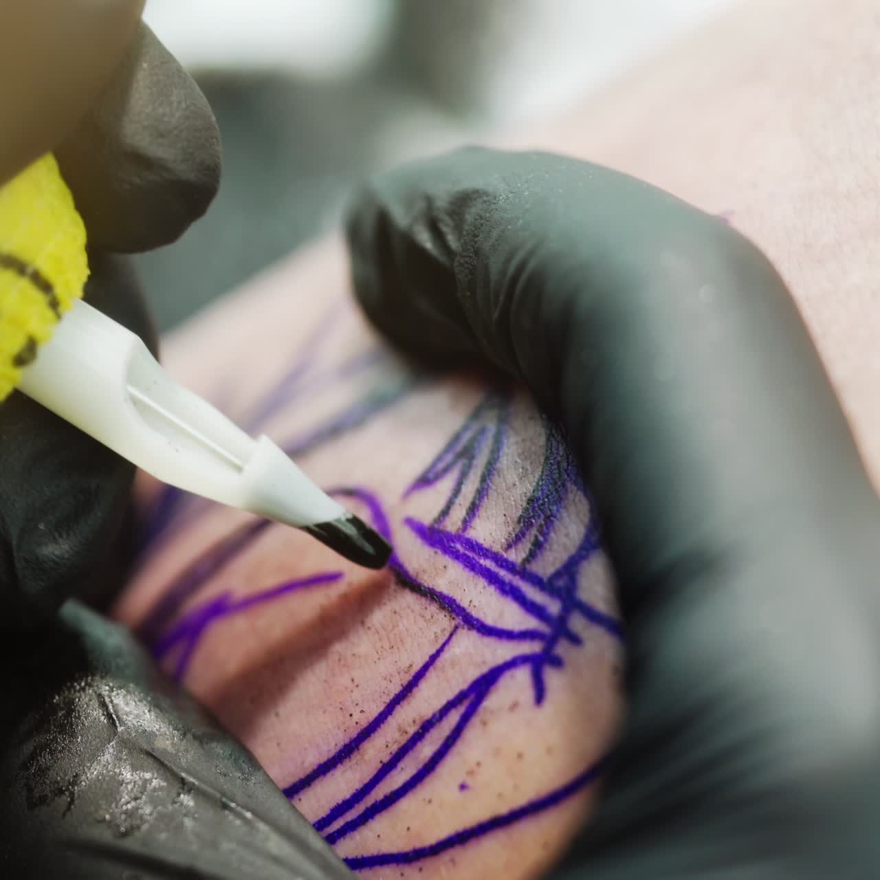 Blue sketched image on skin is covered with black ink from tattoo machine. Gloved hands of tattoo artist stretch the skin white drawing. Close up