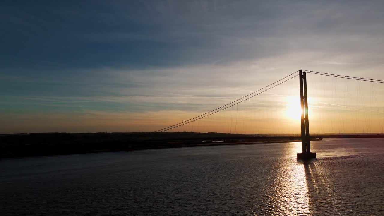 Soaring high above, a drone's eye view captures the Humber Bridge bathed in the warmth of a setting sun, while cars gracefully traverse its span, painting a picture of tranquility in motion