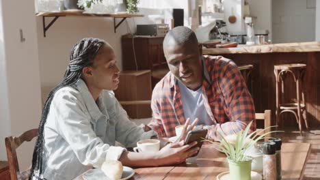 Feliz-Pareja-Afroamericana-Mirando-El-Teléfono-Inteligente-Y-Hablando-En-La-Mesa-En-La-Cafetería,-Cámara-Lenta