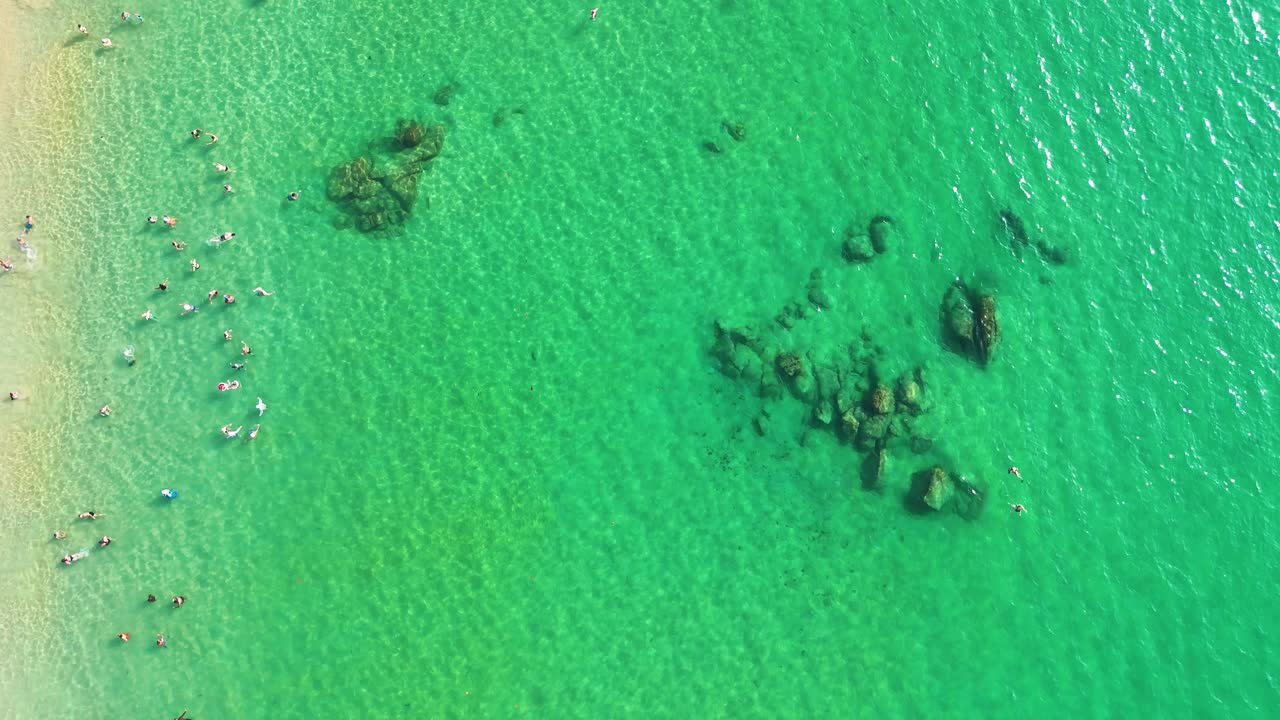 Drone footage captures a crowded Phuket beach with turquoise waters, rocks, and tourists enjoying a sunny day