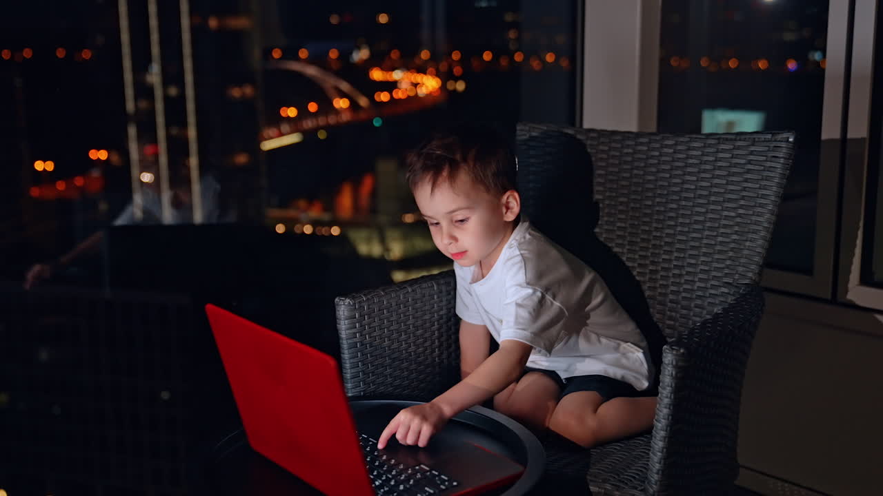 Child using laptop at night in a city. A young child sits in a chair, focused on a laptop screen during the nighttime in a brightly lit urban setting