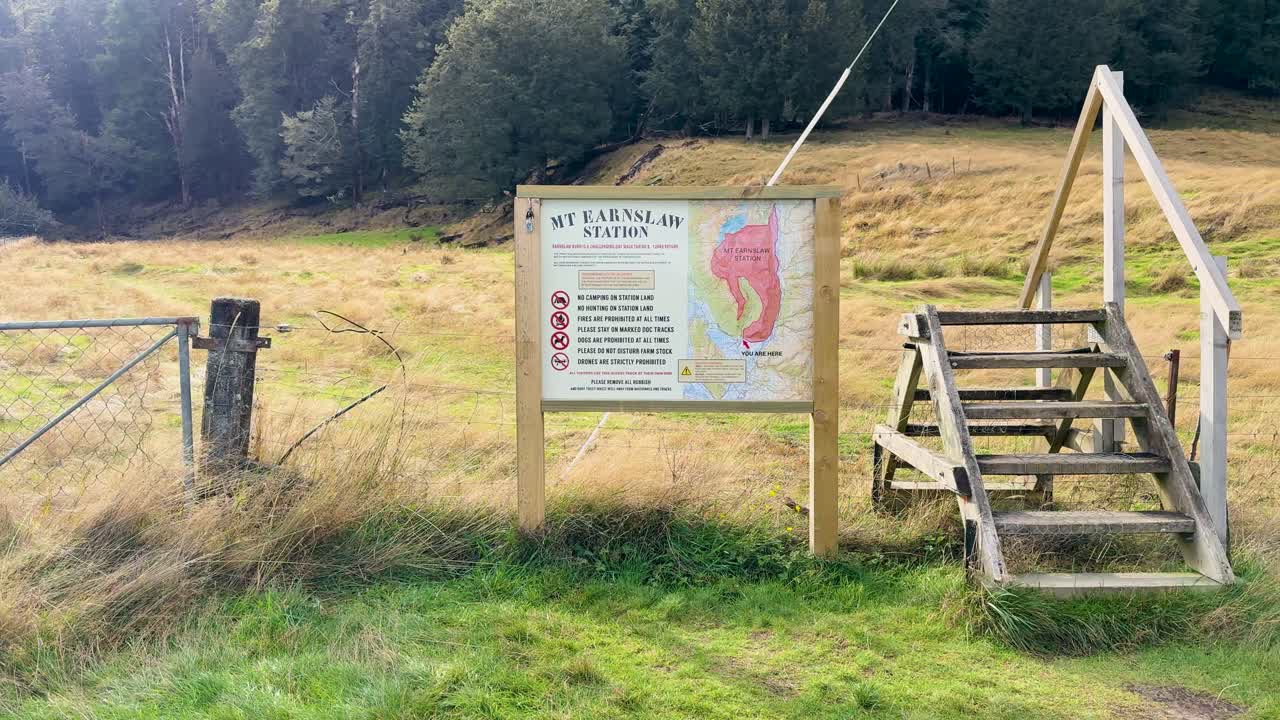 Camera moves toward wooden stile and informational sign in grassy field, natural daylight, steady shot