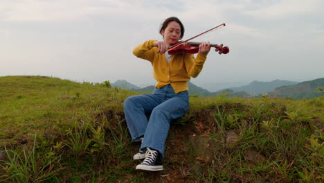 woman peacefully plays the violin outside while perched on an embankment, hair blowing in the wind