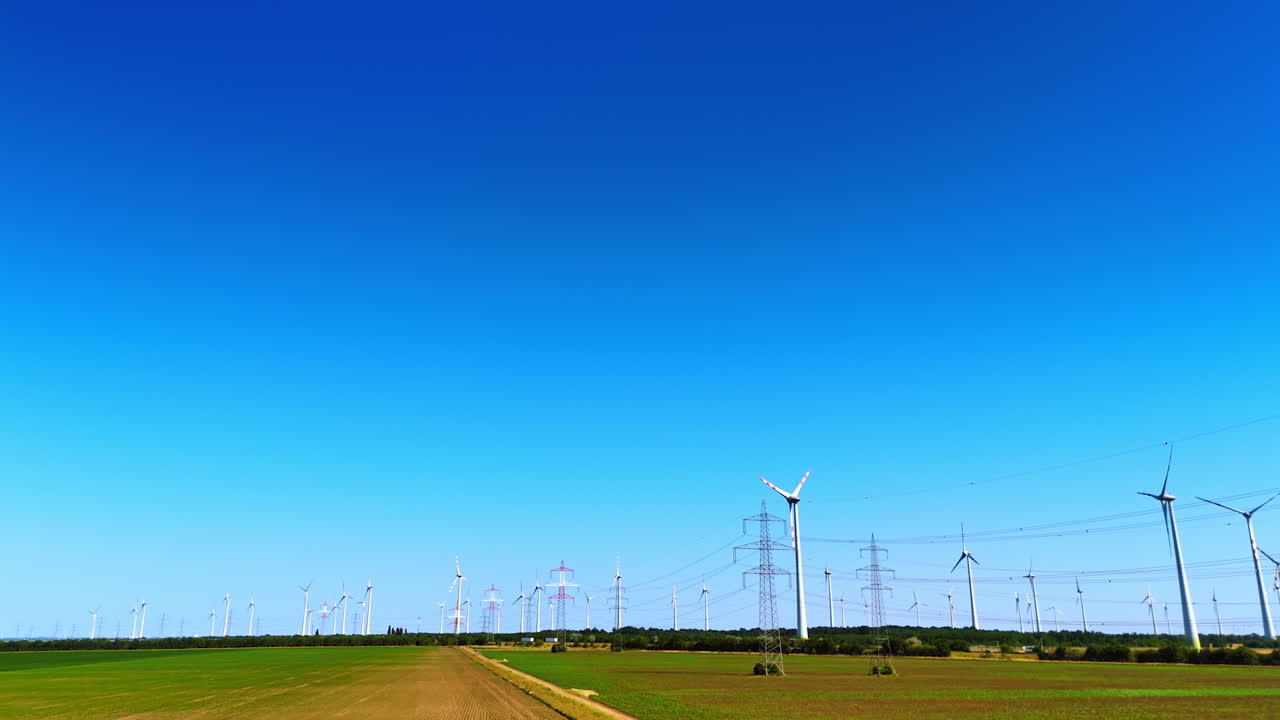 Wind turbines collecting energy. Wind turbines stand tall in a vast green field under a bright blue sky while electric lines stretch across the horizon