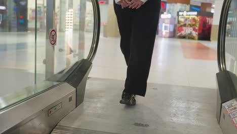 rear view of woman in black pants and dark shoes stepping off moving escalator at top floor inside brightly lit shopping mall with visible storefronts, glass panels, and retail displays in background
