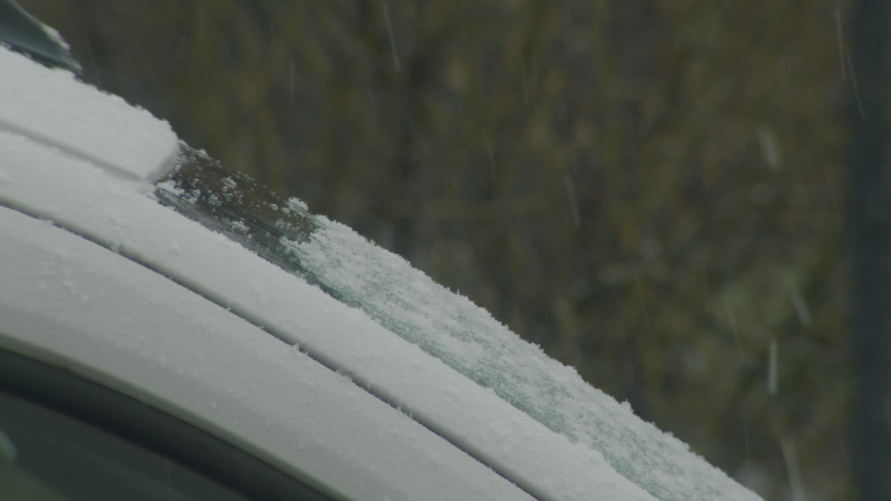 Static close up shot of a cars windshield, view from the side. Snow falling. Windshield covered a little bit.