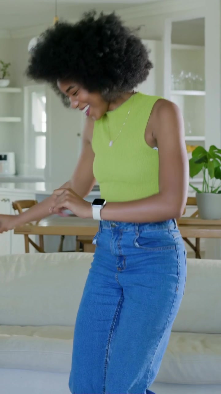 Video verticale: Donna sorridente con top color lime che balla vicino al divano, ride mentre cammina verso la cucina, e un uomo entra.