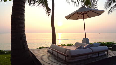 a couchioned couch wedged between palm tree trunks sits empty as the sun sets over a pink ocean horizon