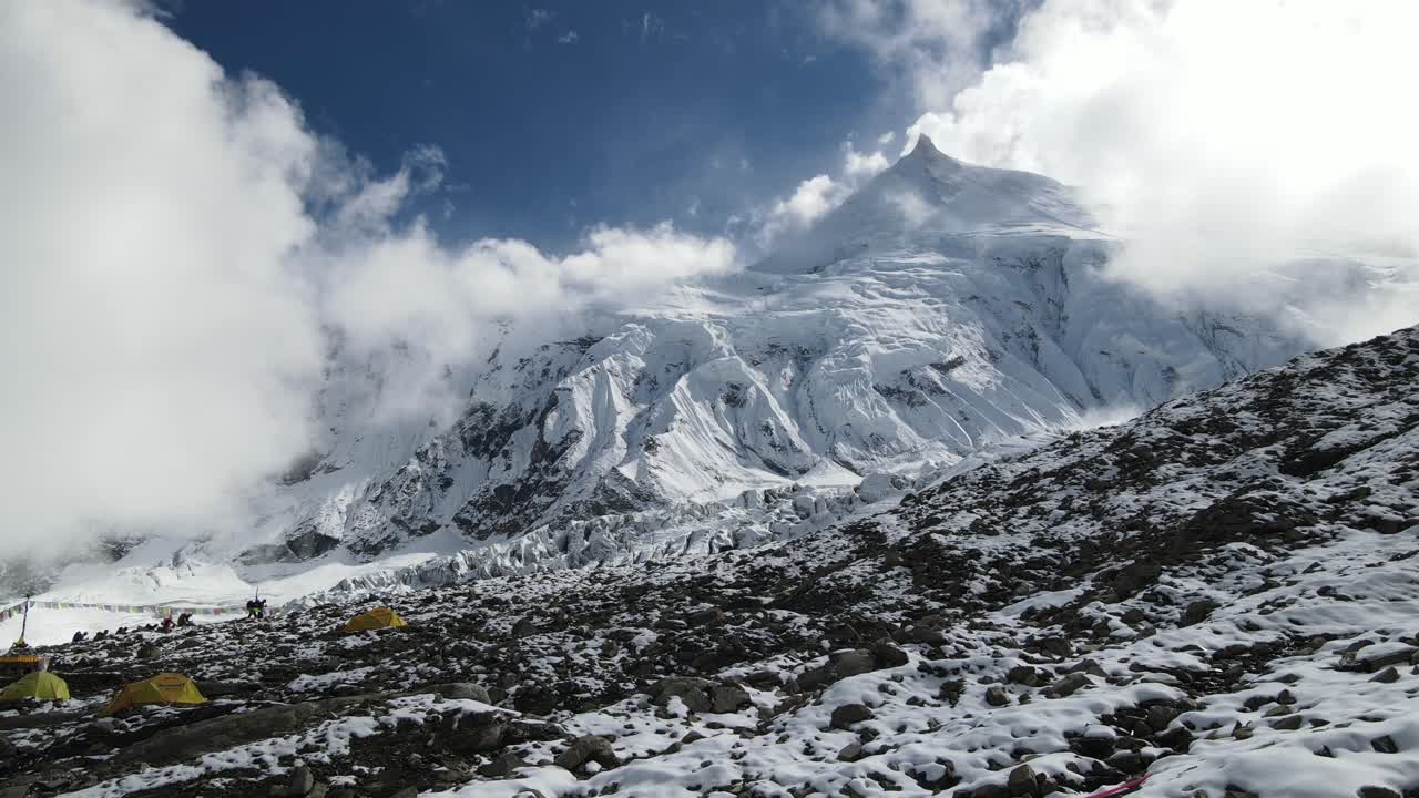 Drone footage showcases Manaslu Base Camp below the 8,163m Himalayan peak. The scene reveals snowy ridges, rugged terrain, and the dramatic high-altitude environment of this remote mountain region