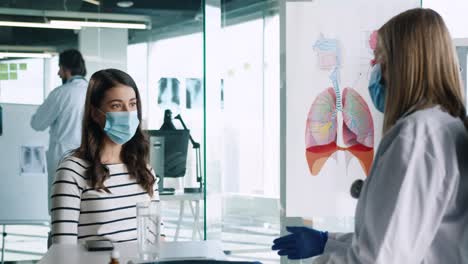 caucasian female doctor wearing medical mask sitting at desk and explaining to female patient treatment for coronavirus in medical consultation