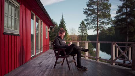 young bearded guy sitting on the porch of cabin while browsing on his mobile phone