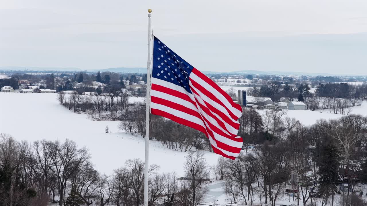 Bandiera americana patriottica che sventola su un pennone davanti a un paesaggio invernale innevato. Ripresa aerea ravvicinata. Campi e case invernali in lontananza. Orgoglio e città fiera degli USA.