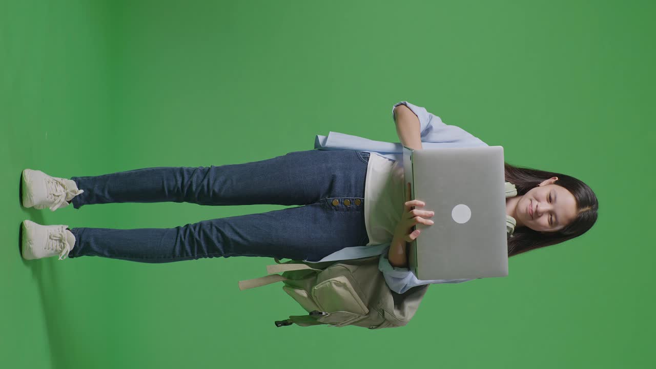 cuerpo lleno de estudiante adolescente asiática con una mochila usar portátil y apuntando a un lado mientras está de pie en el estudio de fondo pantalla verde