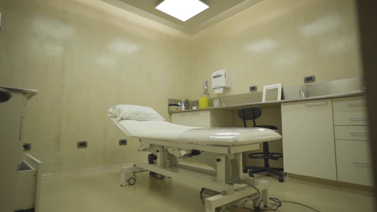 Sala de examen médico vacía con camilla y gabinetes bajo lámpara blanca