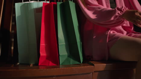 three shopping bags in red, green, and mint green colors are placed on a brown wooden bench by a lady wearing a pink dress, she sits down with her legs crossed, relaxing after shopping