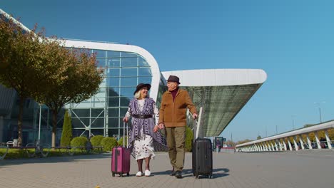 Stylish-retired-family-couple-granny-grandfather-walking-with-luggage-suitcase-bags-to-airport-hall