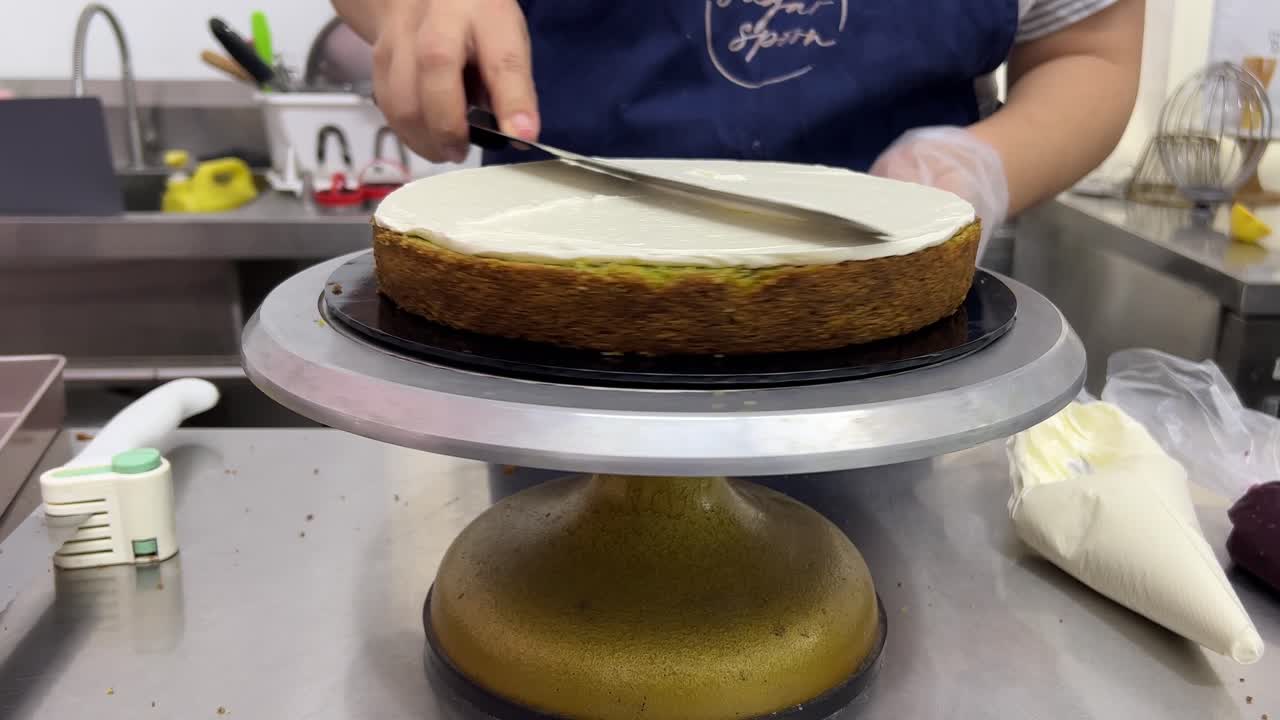 Vista frontale di un pasticcere professionista che liscia la glassa al burro meringata con una spatola, mentre fa ruotare il piatto girevole per torte, in un primo piano ambientato in una pasticceria commerciale.