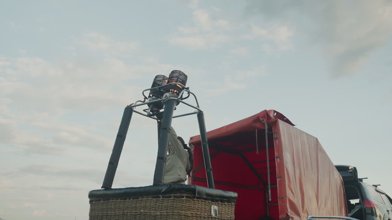woman in light jacket reaches up to ignite hot air balloon burner causing twin flames to roar through metal coils above wicker basket against cloudy sky over grassy field during dusk preparation