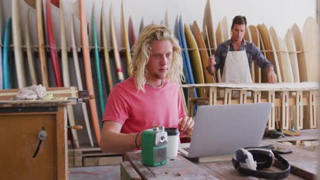 Two-Caucasian-male-surfboard-makers-working-in-their-studio-and-making-a-wooden-surfboard-together